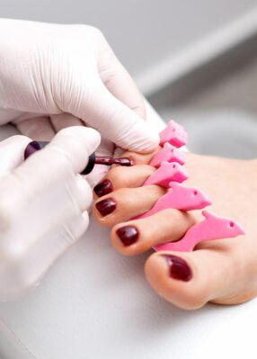 manicure-master-is-painting-female-toenails-with-maroon-nail-polish-by-brush-wearing-white-gloves