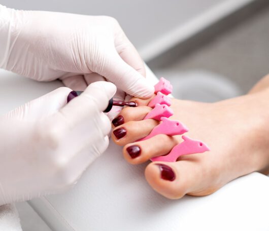 manicure-master-is-painting-female-toenails-with-maroon-nail-polish-by-brush-wearing-white-gloves